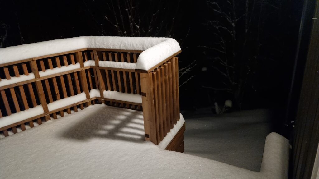 Snow blankets a wooden deck during the night. Surrounding trees are also covered in snow, creating a winter landscape. The scene shows a peaceful outdoor setting.