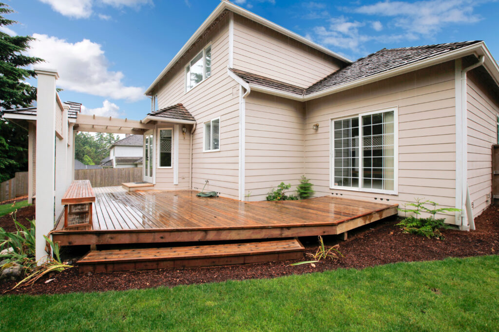 Large beige house with wood deck/porch from the backyard with grass and mulch.
